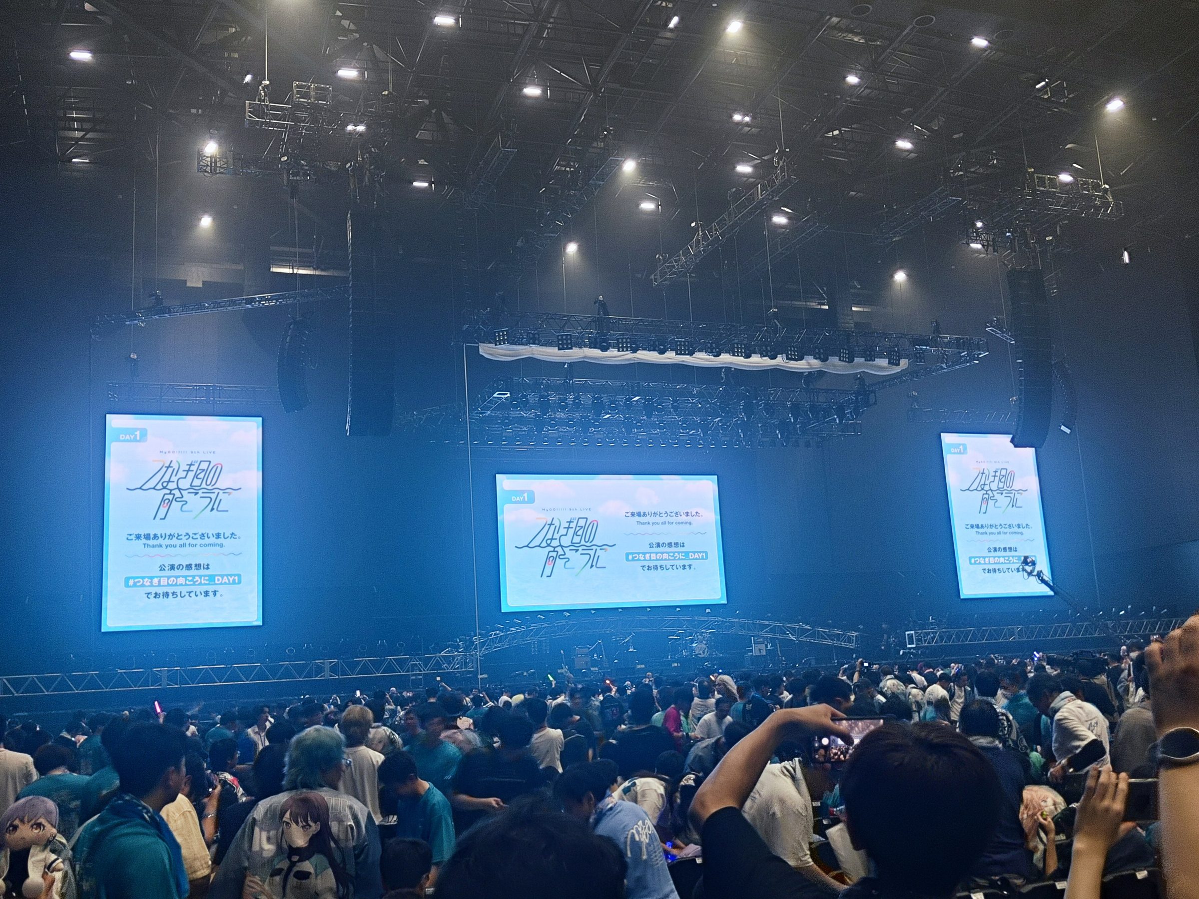 An arena at the end of a concert: two flown curved line-array hangs either side of the stage, overhead lighting rigging, and a full audience below